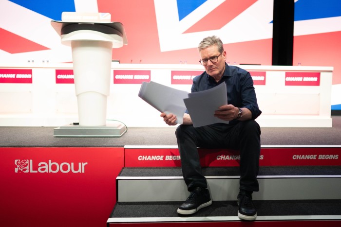 Keir Starmer reheares his speech sitting on steps with Labour slogans and a British flag behind him 