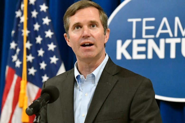 A man in a brown suit speaks into a microphone at a podium. Behind him is an American flag.