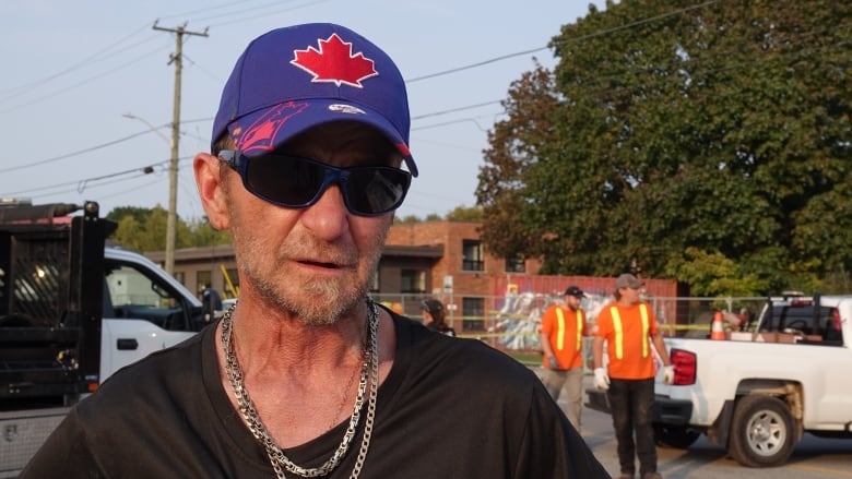 A man in a Blue Jays ball cap and sunglasses stares at the camera. City workers can be seen setting up pylons behind him.