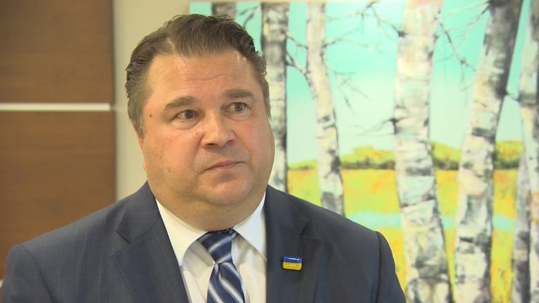 A man in a suit does in interview in front of a backdrop of trees.