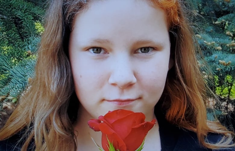 A close-up photo of a teenage girl's face. She has hazel eyes and brown wavy hair, and she's holding a red rose in front of her chin.