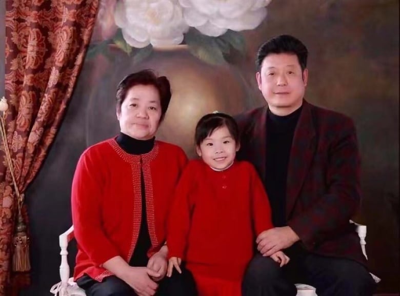 A smiling girl in a red sweater stands between a smiling man and a smiling woman who are sitting on either side of her. They are in a photo studio.  