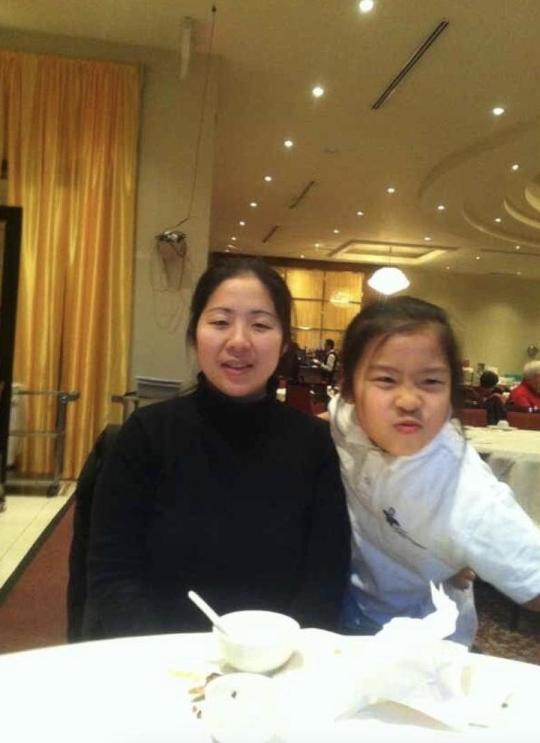 A girl makes a funny face while standing next to a woman sitting in a restaurant. 