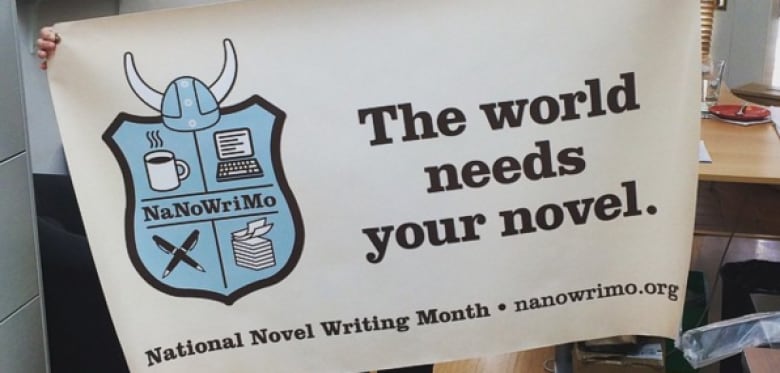 A woman holds up a poster that reads "the world needs your novel - National Novel Writing Month - nanowrimo.org