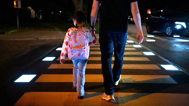 A young girl with a backpack and a man in jeans walk across a crosswalk at night, with lights lining the sides of the crosswalk