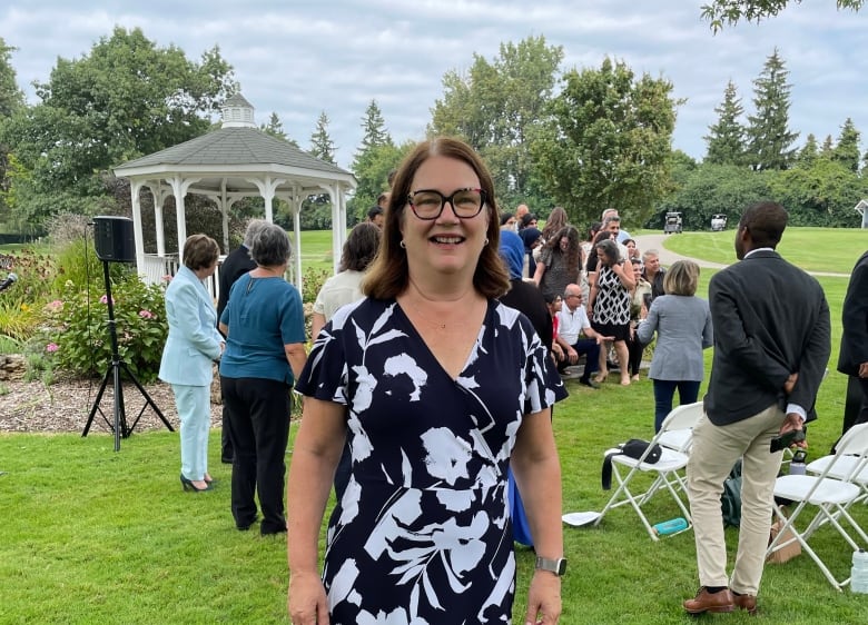 A woman in a black and white floral patterned dress poses on a lawn where other people, chairs and a pergola are seen in the background as an event unfolds.