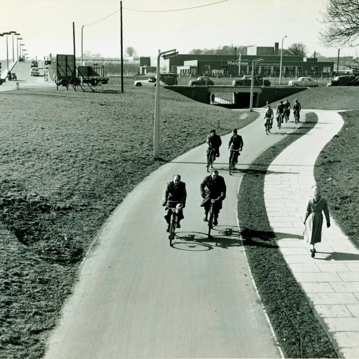 A black and white photo from the late 1950s or early 1960s showing a sunlit view of cyclists on a cycle path and pedestrians on a footpath, well away from the motor traffic in the background