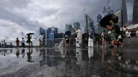 Tourists are posing and walking at Merlion Park in Singapore with the central business district skyline in the background. Some people are holding umbrellas, and the ground is wet, reflecting their figures. The iconic Merlion statue is visible among the tourists.