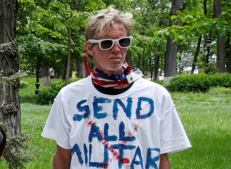 A man wearing white-framed sunglasses and a tshirt, with stubble on his face, is shown in an outdoor photo.