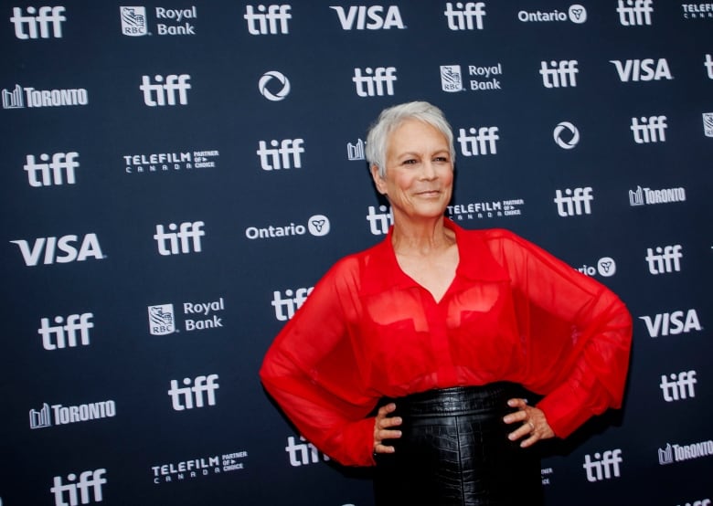 A woman with grey hair, wearing a red blouse and a black skirt, poses for a photo in front of a dark blue backdrop with the words "tiff" and Visa" printed on it.