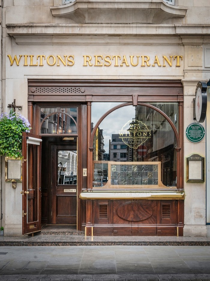 The exterior of Wiltons Restaurant, featuring a classic wooden facade with arched windows and gold lettering on the stone wall above the entrance