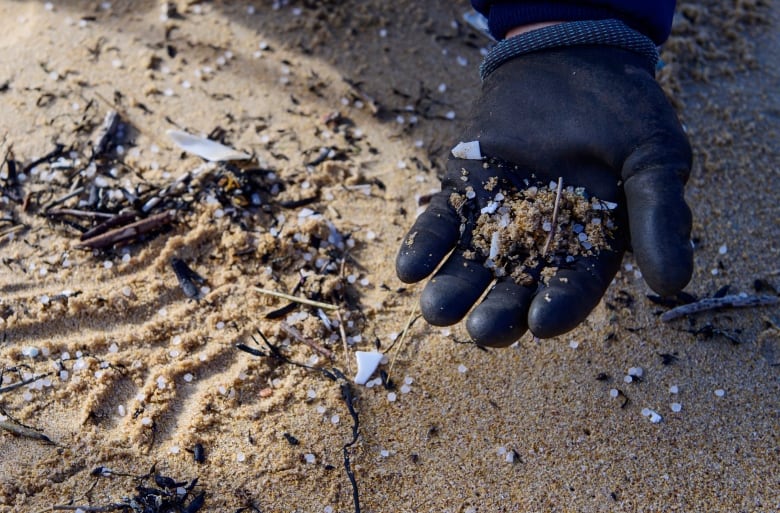 A hand wearing a black glove holds clumps of sand and plastic, with sand seen in the background filled with more plastic.