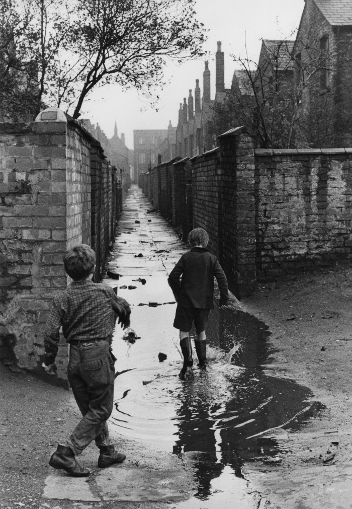 Black-and-white picture of two young children playing in a flooded urban alleyway in Manchester in the 1960s