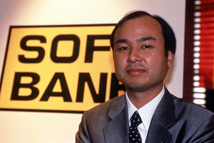 A smiling man in his forties, in suit jacket and tie. Behind him is a yellow sign with black lettering saying SoftBank