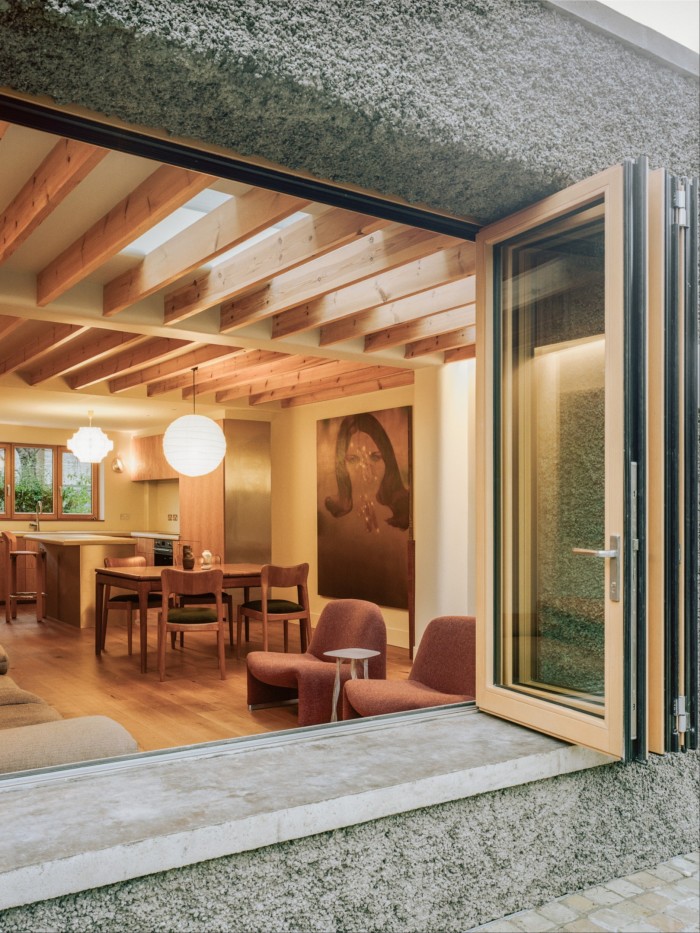 A kitchen, photographed from the outside through a large open sliding window. A large modern painting of a woman hangs on one wall; the ceiling has wooden beams, and the flooring and dining furniture are also wooden