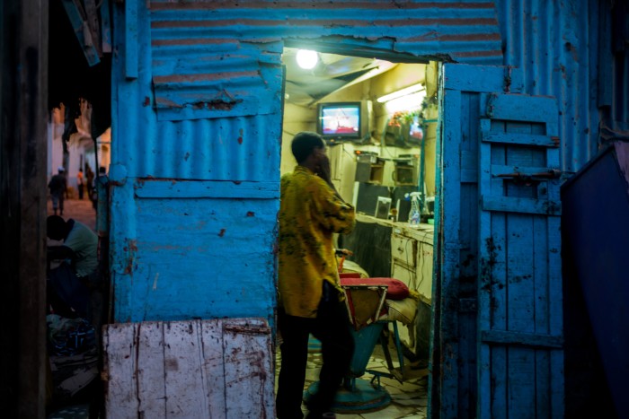 A person in a yellow shirt standing inside a small, rustic barbershop with blue corrugated metal walls, watching a television mounted on the wall