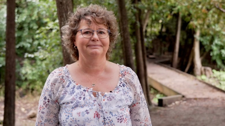 A woman in a floral blouse smiles at the camera while standing in a bright forest clearing, a pathway seen behind her.