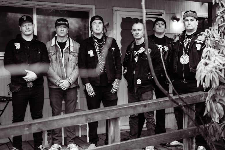 Six men in dark clothing stand outside, looking at the camera and posing on the porch of a house. The photo is in black and white.