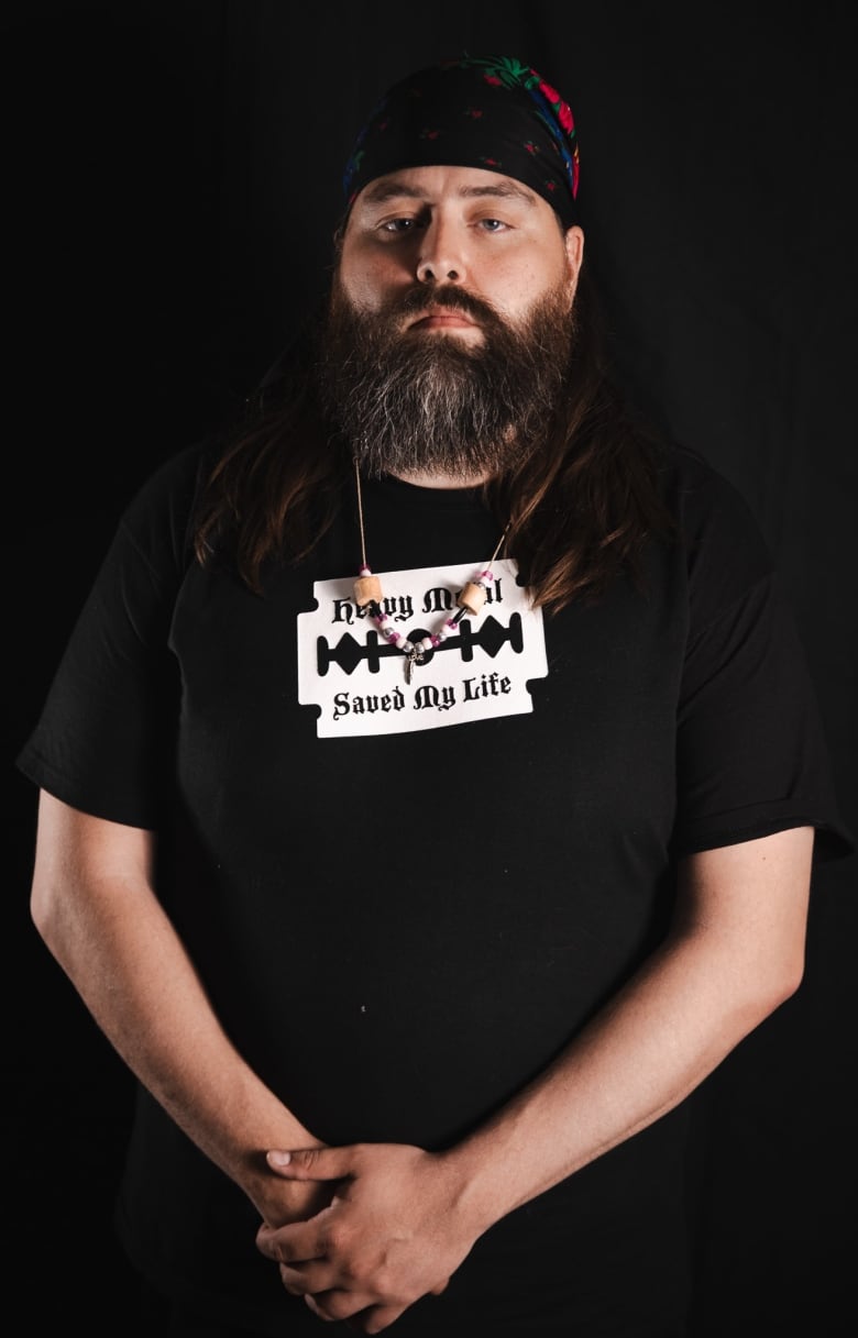 A man in a dark t-shirt and a bandana looks at the camera with a serious face. His hands are folded in front of him.