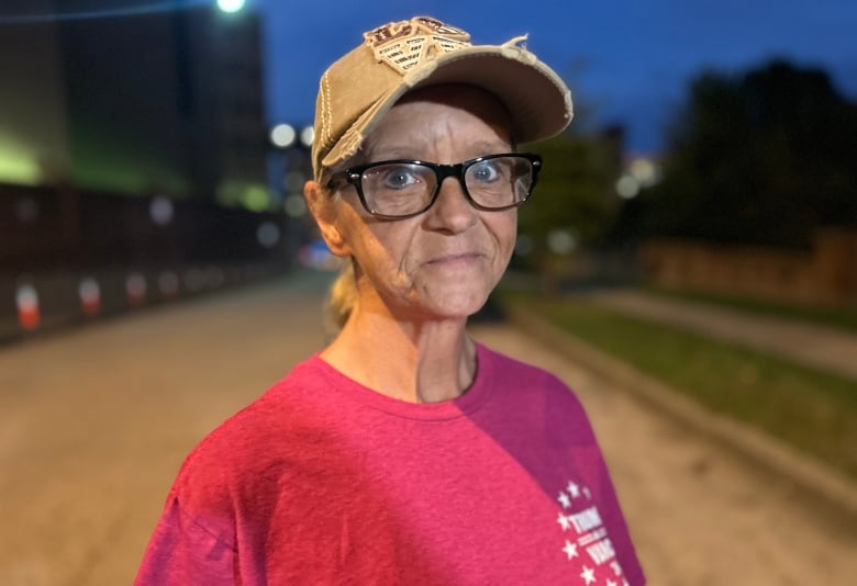 A woman stands outside at night, looking at the camera. She's wearing a pink T-shirt, black rimmed glasses and a baseball cap with 'love' written on it.