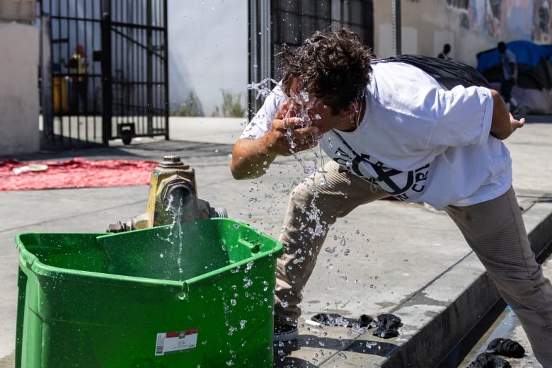 A guy in a white t-shirt is splashing himself in the face and drinking some of the water from a fire hydrant on a hot California street.