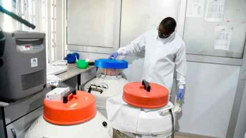 An individual wearing a white protective lab coat, gloves, and glasses is opening what seems to be a cryogenic tank