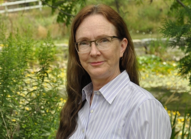 An adult woman with long red-brown hair wearing classes and a pale blue-grey shirt poses for a photo outside in front of trees and a creek.