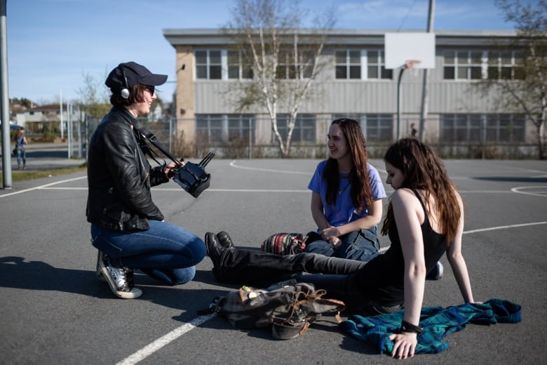 A woman with a camera talks to two teenage girls sitting in a parking lot. 