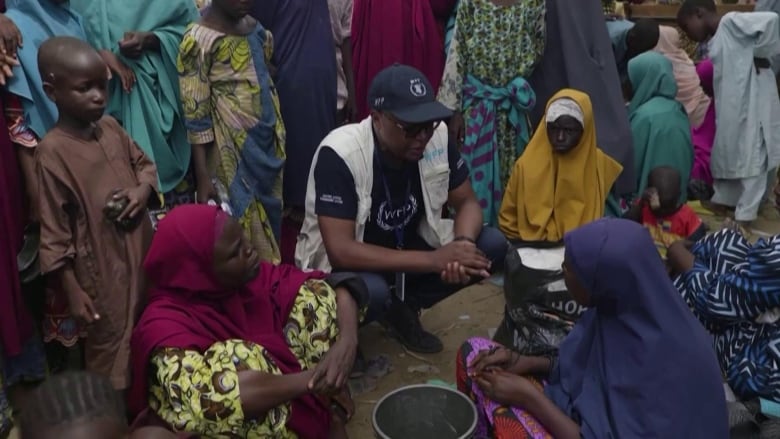 A man in a World Food Program hat and vest kneels among a group of women and children.