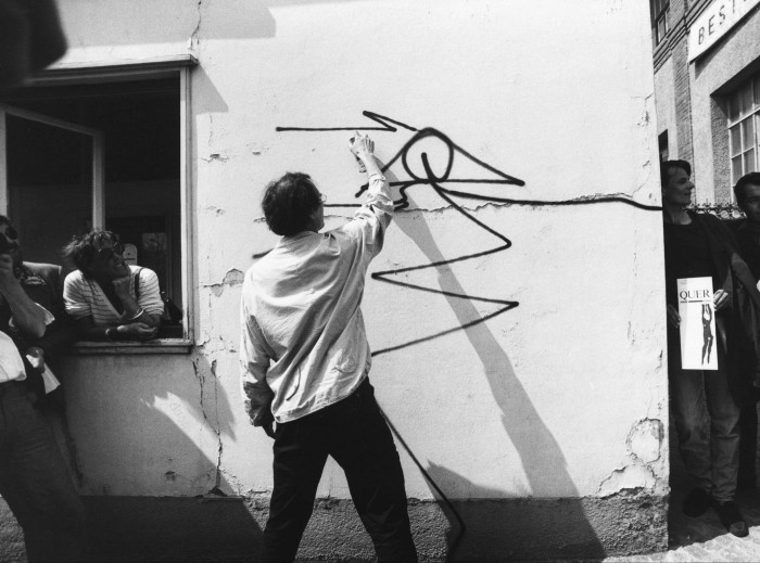 A black and white photograph of street artist Harald Naegeli spray-painting a work onto the wall of a building in the late 1970s