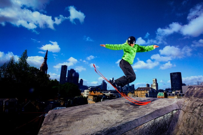 A woman in bright green jacket, dark trousers and black helmet performing a ski stunt on a dry  slope against a cityscape