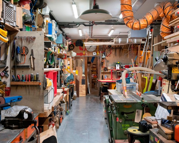 A workshop with grey floor and strip lighting is filled floor-to-ceiling with tools, material and objects, from screwdrivers and saws, to stacks of pieces of wood, chair frames and a garden gnome 