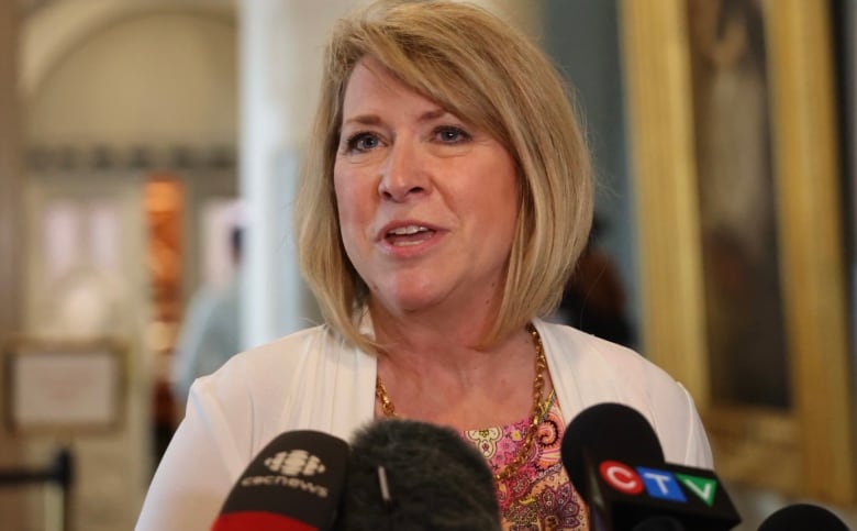 A woman with short blond hair speaks into multiple microphones.  