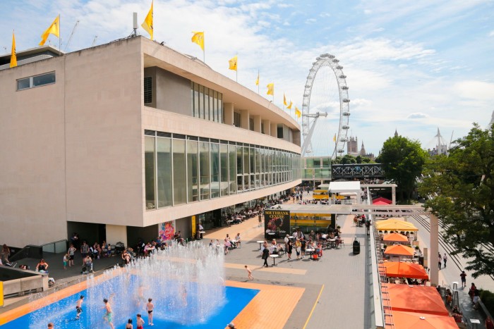 An angled view of a large concrete and glass building. The London Eye can be seen in the background, and children are splashing in fountains in the foreground. A row of cafe seating areas with orange canopies is in a row in front of the building