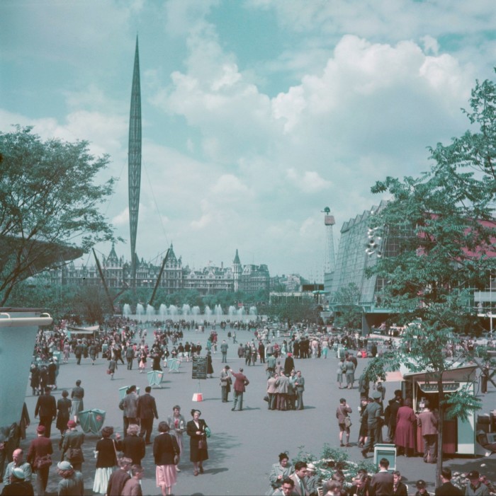 An old colour photograph of the south bank of the river Thames, taken during the Festival of Britain. Crowds of people walk there, and the tall, vertical structure of the Skylon appears to be almost floating in the middle distance 