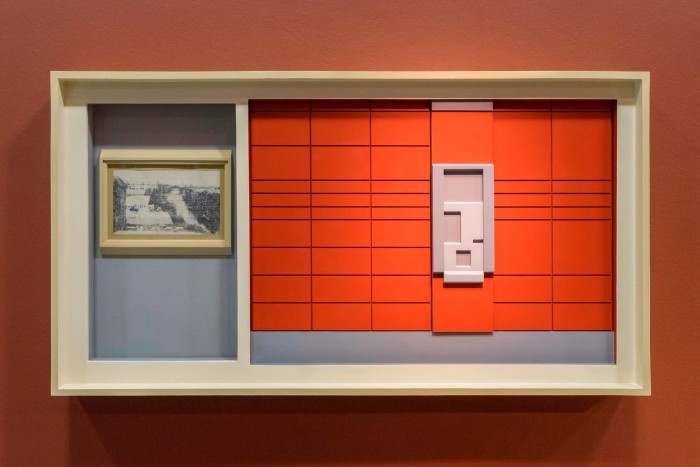 A framed, geometric three-dimensional work made from various types of industrial wood, cut into rectangular shapes, coloured orange and grey and resembling a piece of stationary or a computer floppy disk 