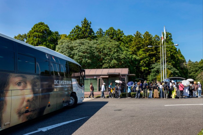 A queue of people forms after disembarking from a museum bus, its side emblazoned with a large portrait of an Old Master