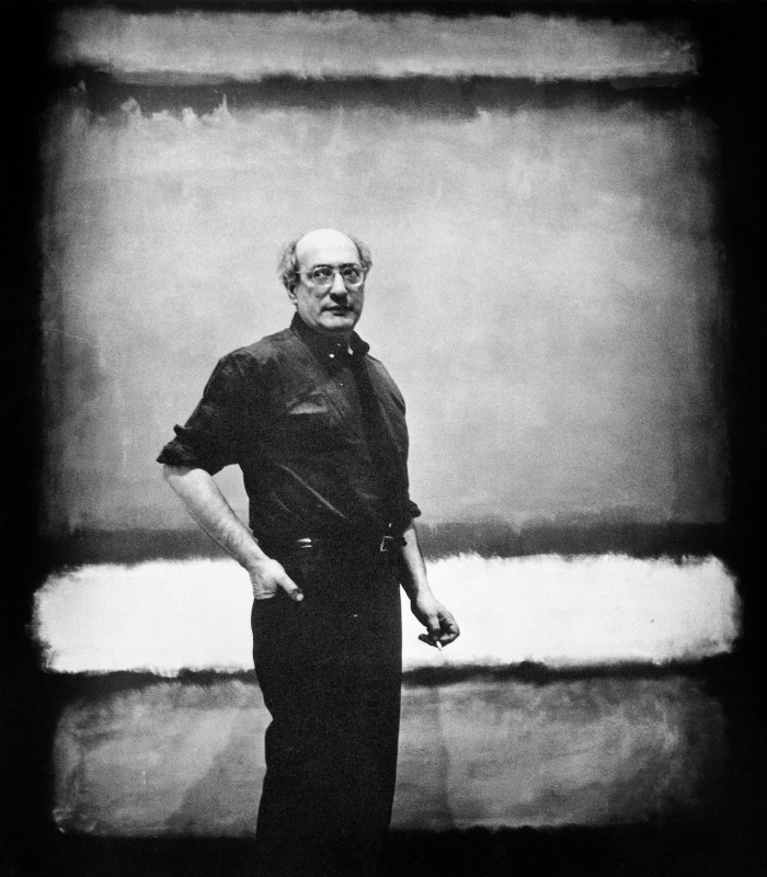 A black and white photo of a bald man standing in front of large painting 