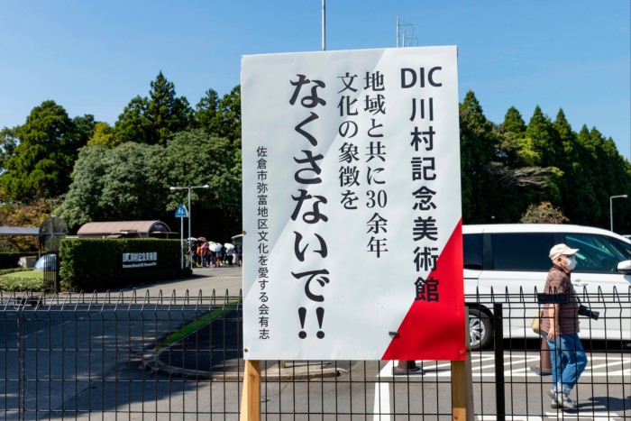 A large white sign with Japanese writing on it. The sign is attached to railings. There is a white car and a person in the background