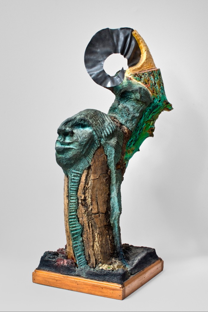 Sculpture of what looks like a tree stump overlaid with a face and tale, made from a dark green material that looks like tarnished copper