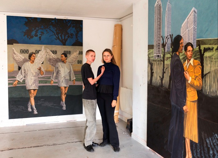 Two women in an art studio, standing in front of colourful paintings of people in front of urban tower blocks