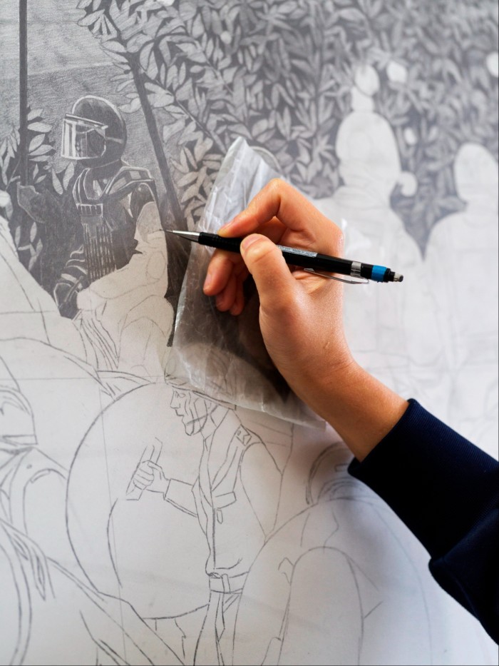 close up of a hand drawing with a mechanical pencil, working on a detailed black-and-white drawing which includes a portrait of a riot cop 