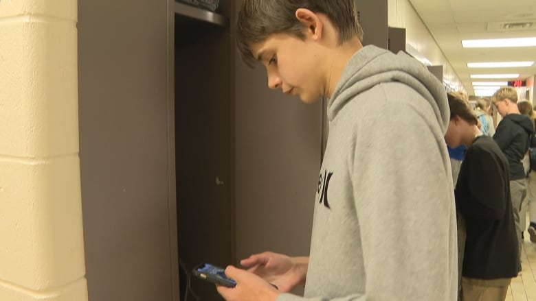 A teen boy in a grey hooded sweatshirt looks down at a cellphone while standing in front of an open locker, with other students seen in a school hallway behind him.