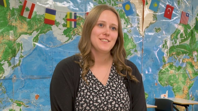 A female teacher in a dark floral dress and black cardigan stands in a classroom, with a large map of the world and a string of flags seen on display behind her.