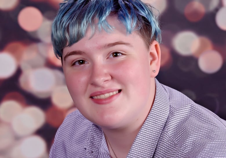 A portrait of a teen with a short-cropped blue haircut smiling, with twinkling lights blurred in the background.