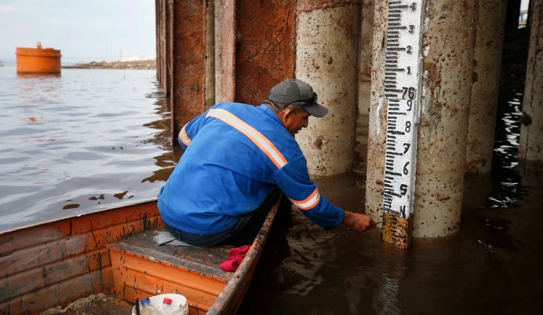 A man in a blue sweater sits in a rowboat floating in water next to tall metal posts that vanish into the top of the image. He is leaning towards one, which has a depth marker on it with numbers showing the depth of the water. 