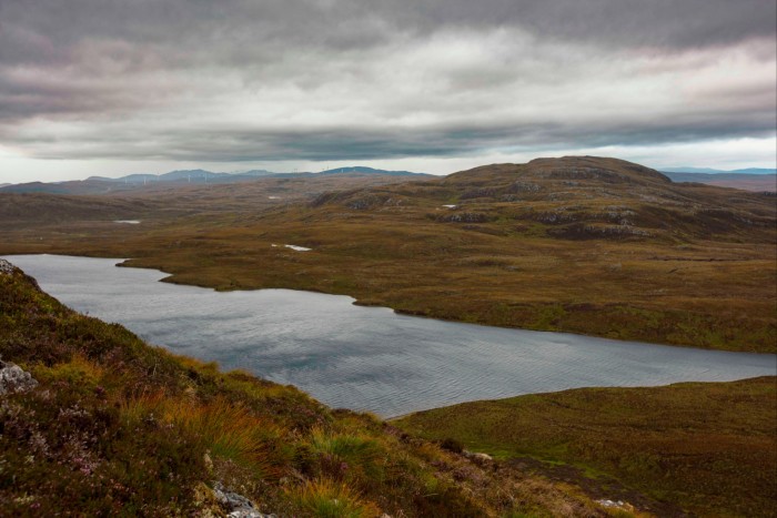 A panoramic view of Loch nam Breac Dearga under a cloudy sky. The landscape features rolling hills covered with brown and green vegetation, surrounding the expansive, calm waters of the loch