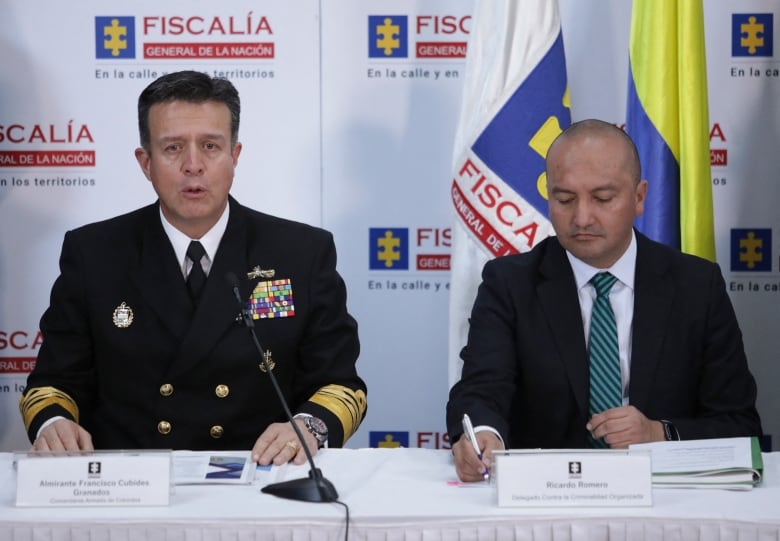 A man in a navy uniform and a man in a suit sit at a table in front of a microphone. 