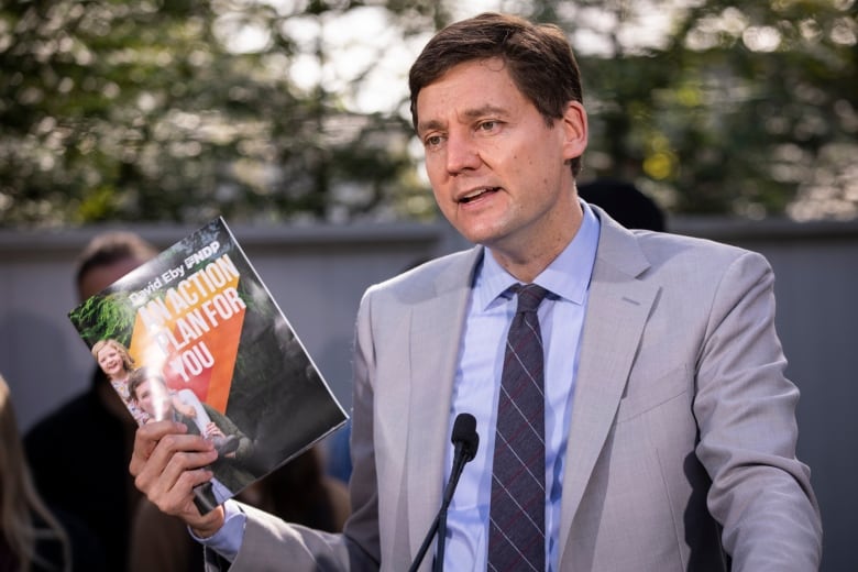 A man holds up a pamphlet reading 'David Eby BC NDP An Action Plan For You'.