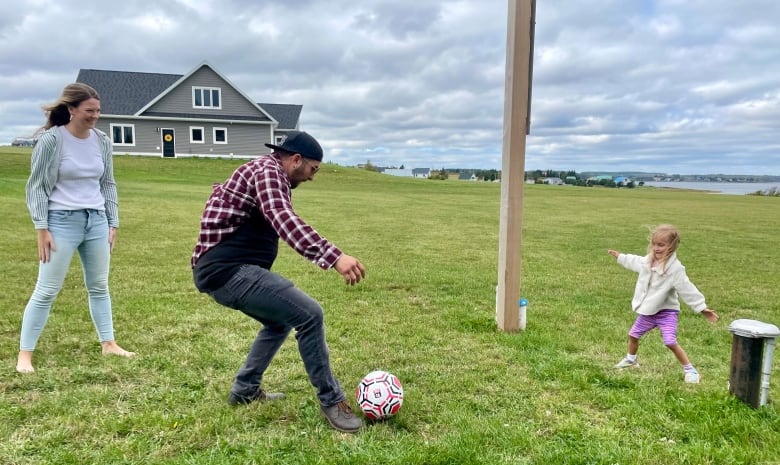 Little blond girl with adult man and woman kicks soccer ball around lawn by the seaside home